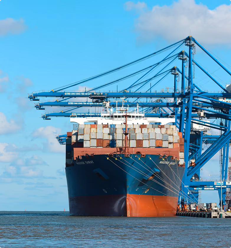 Front view of a large cargo ship loaded with shipping containers docked at a port, with several blue cranes extended over the ship against a partly cloudy blue sky.