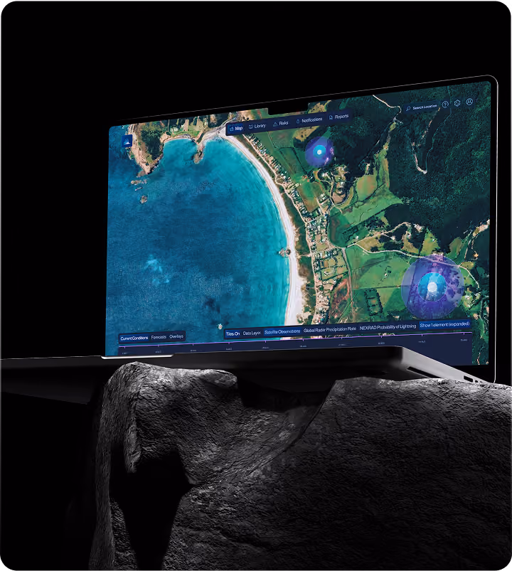Laptop resting on a dark rock surface displaying a satellite map view of a coastal area with blue water, a sandy beach, green land with buildings, and overlay icons indicating data points or observations.