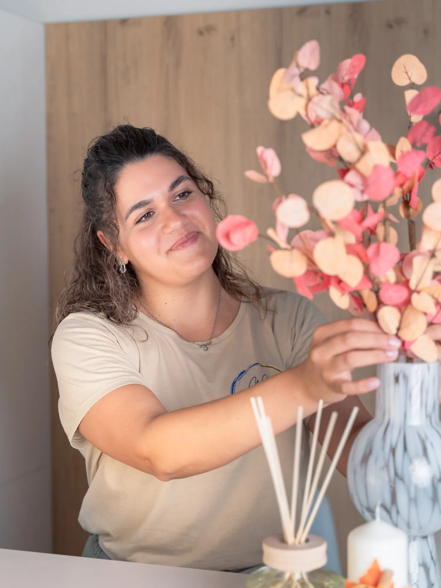 A woman with curly dark hair wearing a beige t-shirt arranges pink and peach dried eucalyptus stems in a marbled gray vase on a table with a reed diffuser and candle.