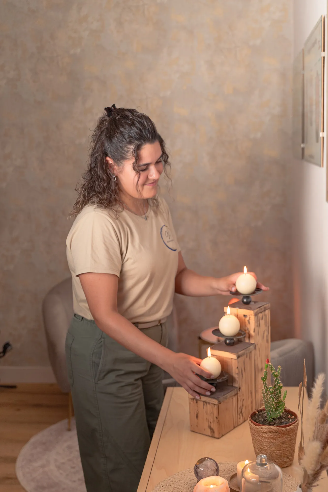 Woman with curly hair in a beige t-shirt and green pants lighting or arranging three lit round candles placed on a tiered wooden stand on a table with a potted cactus and other decorative items in a cozy room.