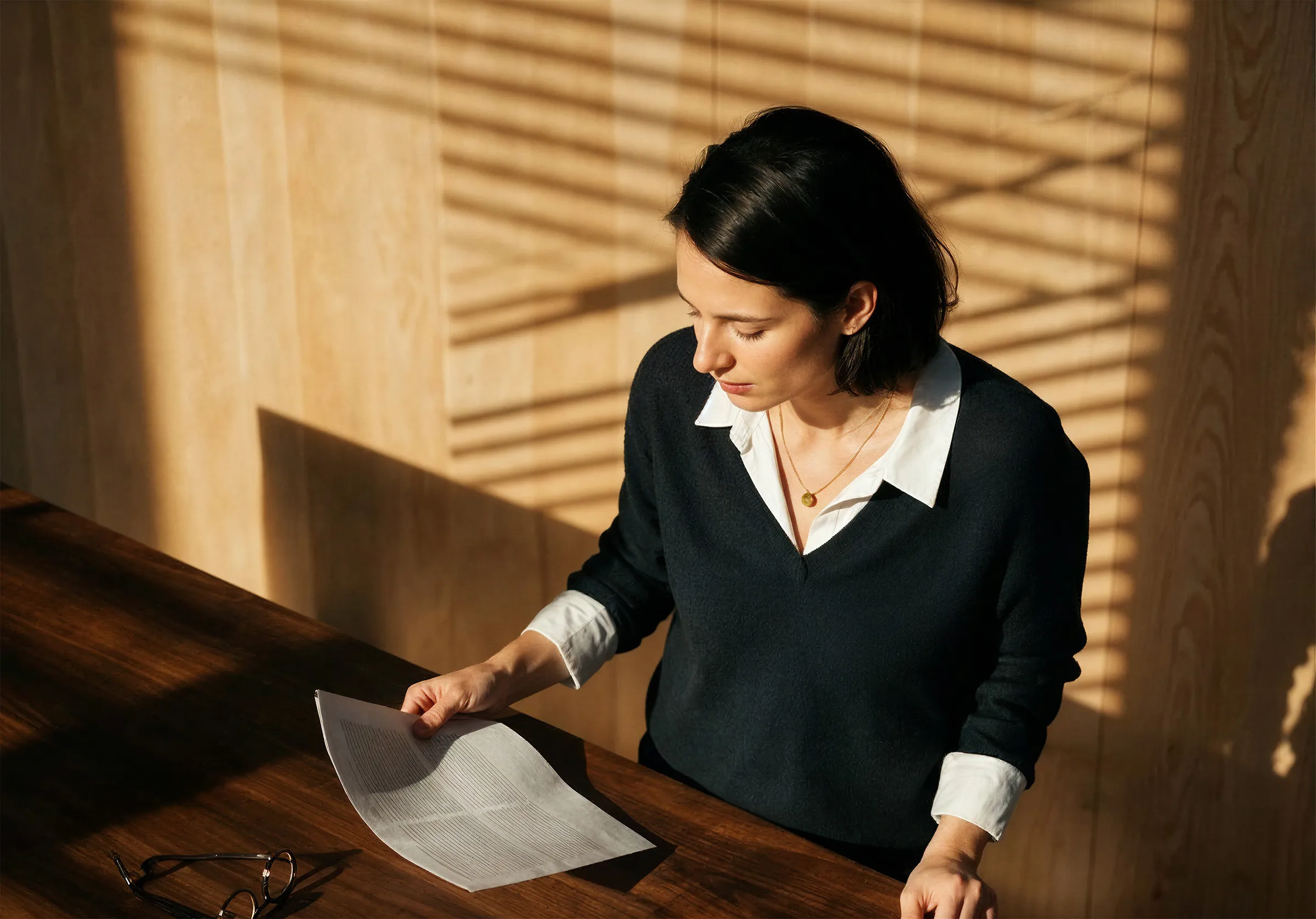 Woman with dark hair wearing a black sweater and white shirt reading a paper at a wooden table with eyeglasses on it in sunlight.