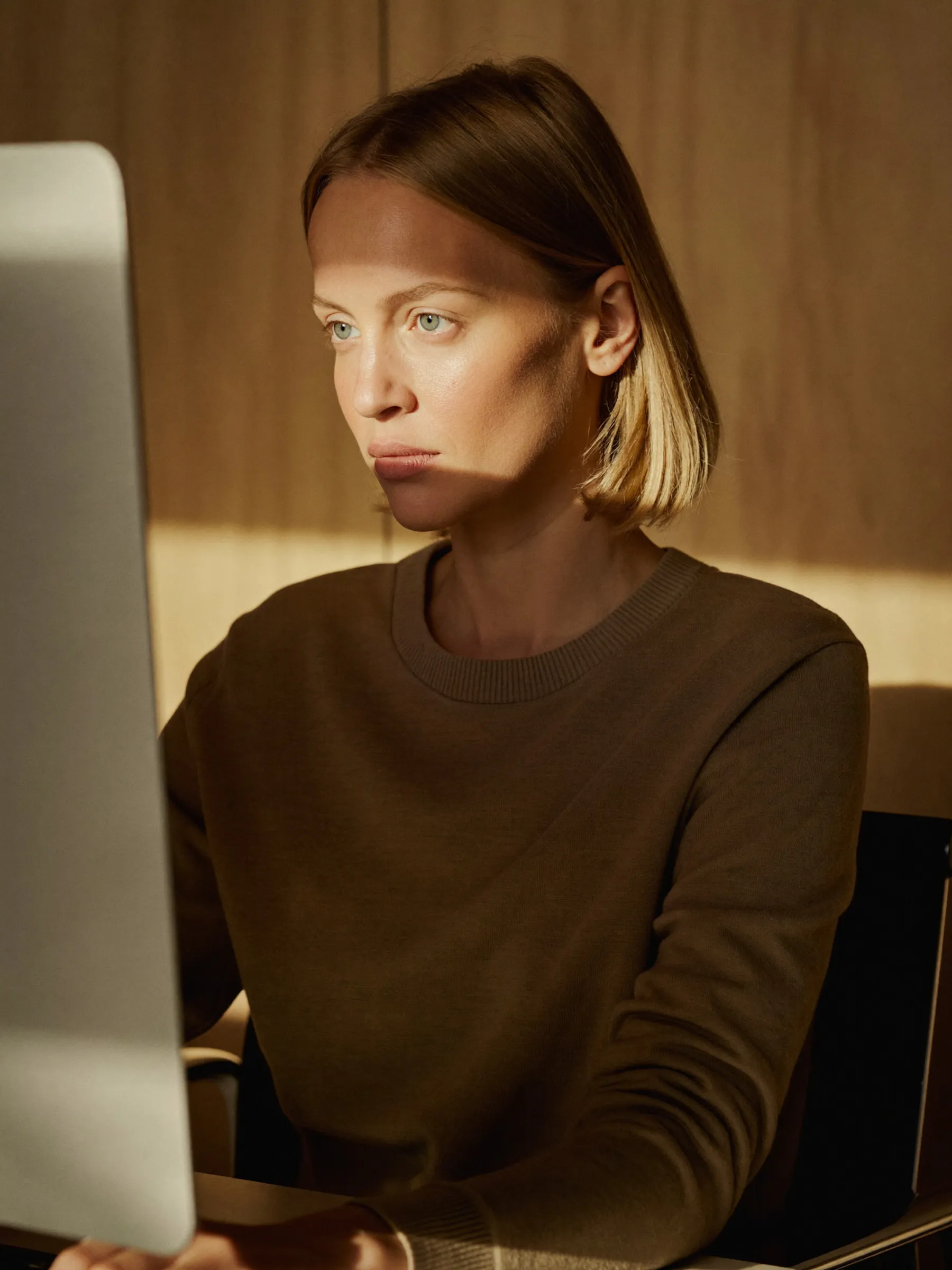 Focused woman with blonde hair working on a computer in warm natural light indoors.