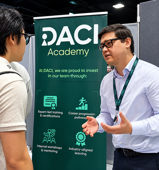 Two men in glasses discussing in front of a DACI Academy banner about team investment programs.