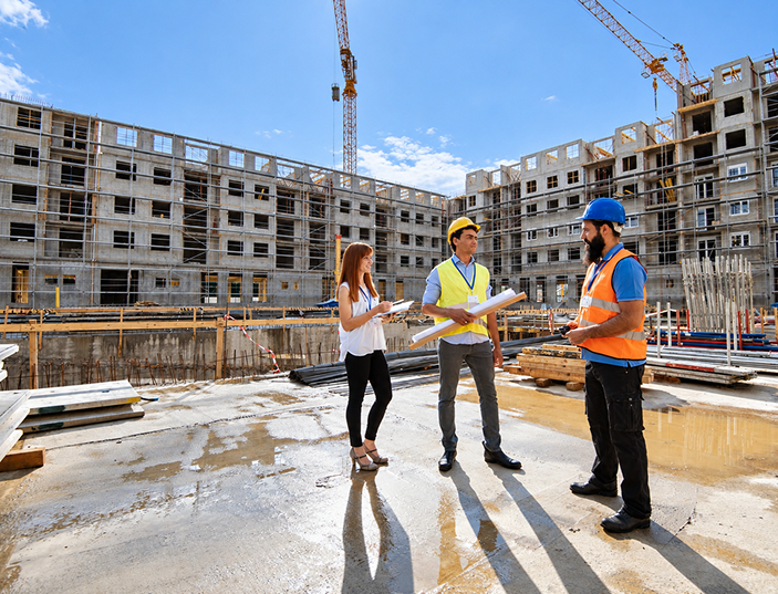 Three construction professionals wearing safety gear discuss plans at a building site on a sunny day.