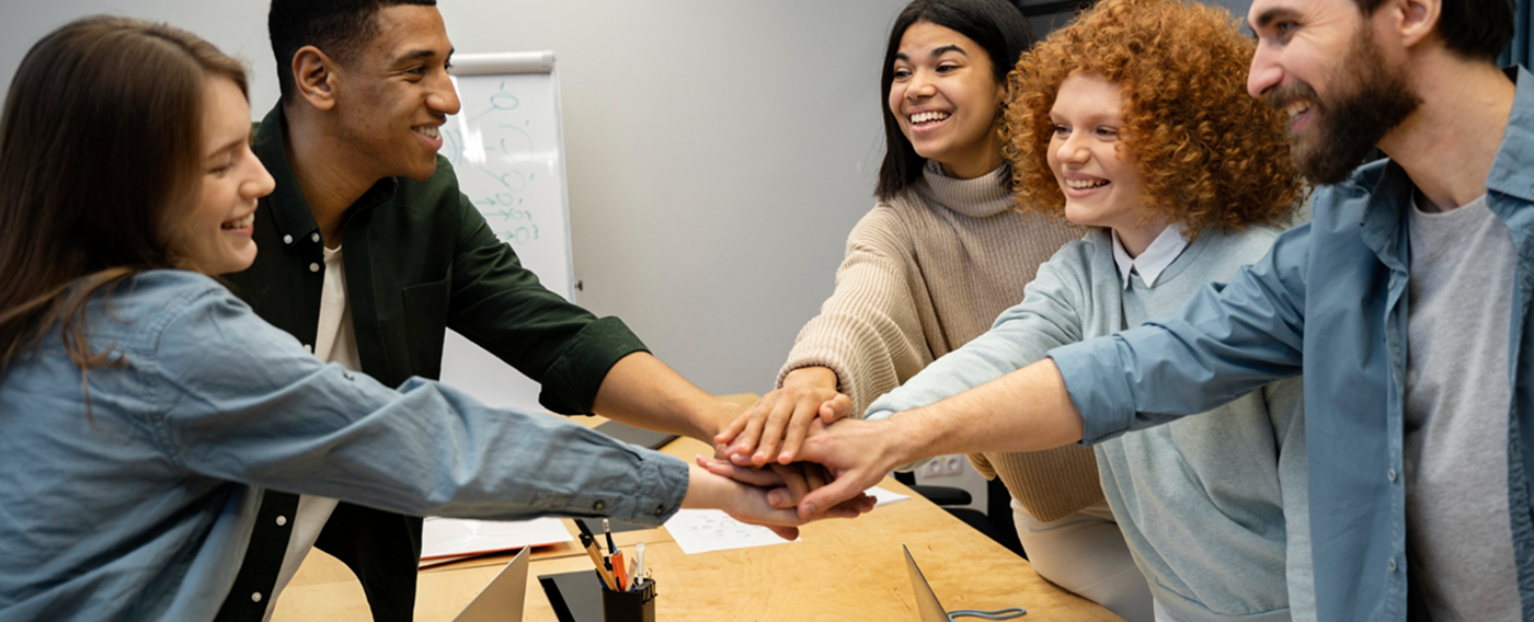 Five diverse coworkers smiling and stacking hands in a team gesture over a desk in an office.