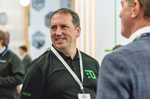Man in a black polo shirt with a white lanyard smiling and talking to another man in a blue jacket in an indoor setting with blurred people and signs in the background.