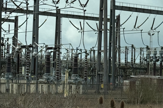 Electrical substation with numerous insulators, wires, and metal framework, surrounded by a fence and some dried vegetation in the foreground.