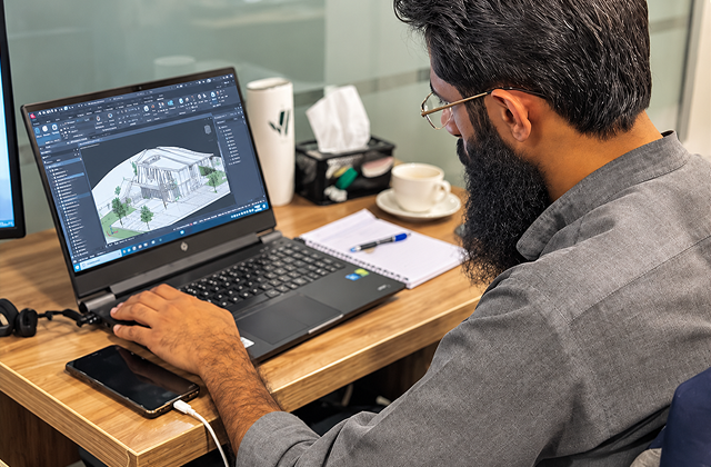 Man with glasses and beard working on architectural 3D building design on a laptop at a wooden desk with notebook, coffee cup, and tissues.