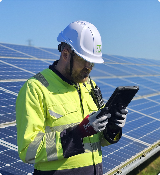 Worker in a high-visibility jacket and white hard hat using a tablet near solar panels under a clear blue sky.