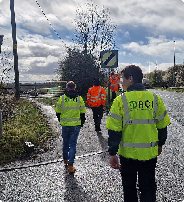 Four people wearing high-visibility jackets walk along a roadside path; two jackets show 'DACI' branding, and the sky is partly cloudy with bare trees nearby.