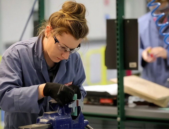 Woman wearing safety glasses and gloves focused on working with a vise clamp in an industrial or workshop setting.