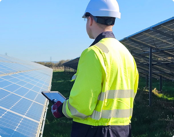 Engineer in a hi-vis jacket and hard hat using a tablet beside rows of solar panels.