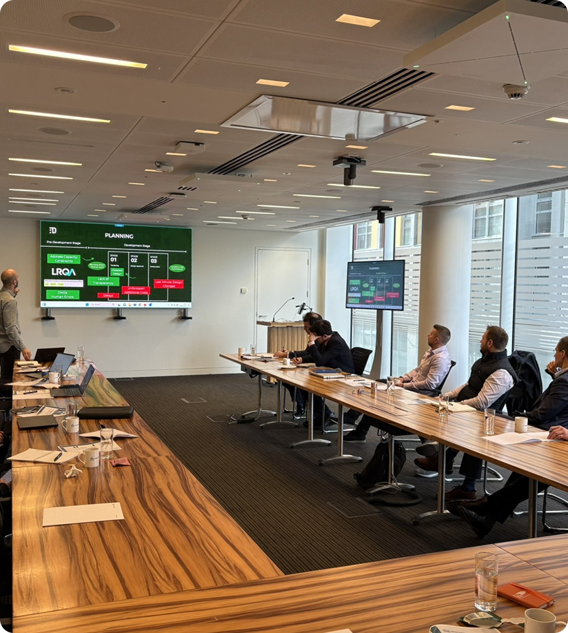 Conference room with a group of seven people seated around wooden tables listening to a presenter standing beside a large screen showing a planning presentation with development stages and key points in green and red boxes.