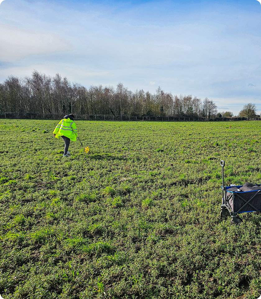 Person wearing a neon yellow reflective jacket using a measuring wheel in a green field with sparse grass during daytime, trees in the background, and a folded wagon cart on the right side.