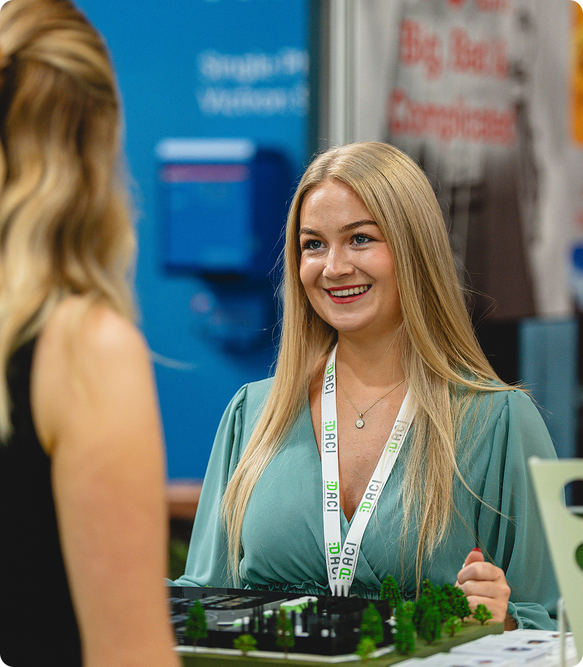 Smiling woman with long blonde hair wearing a green blouse and a white DACI lanyard talks to another person, with a miniature model of a building and trees in front of her.