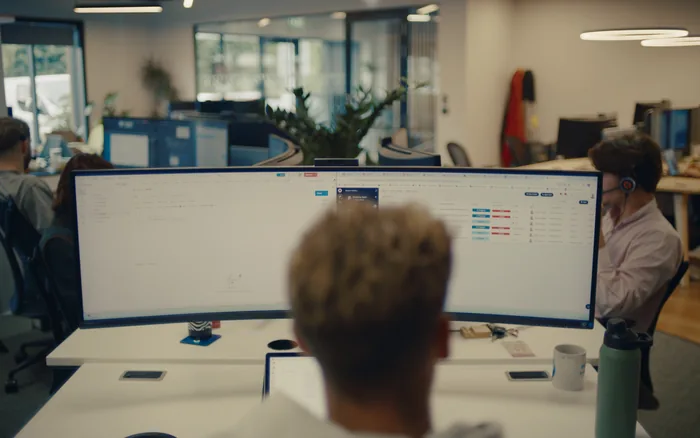 A Halo team member working through a support ticket queue on a wide monitor, with a colleague on a headset nearby
