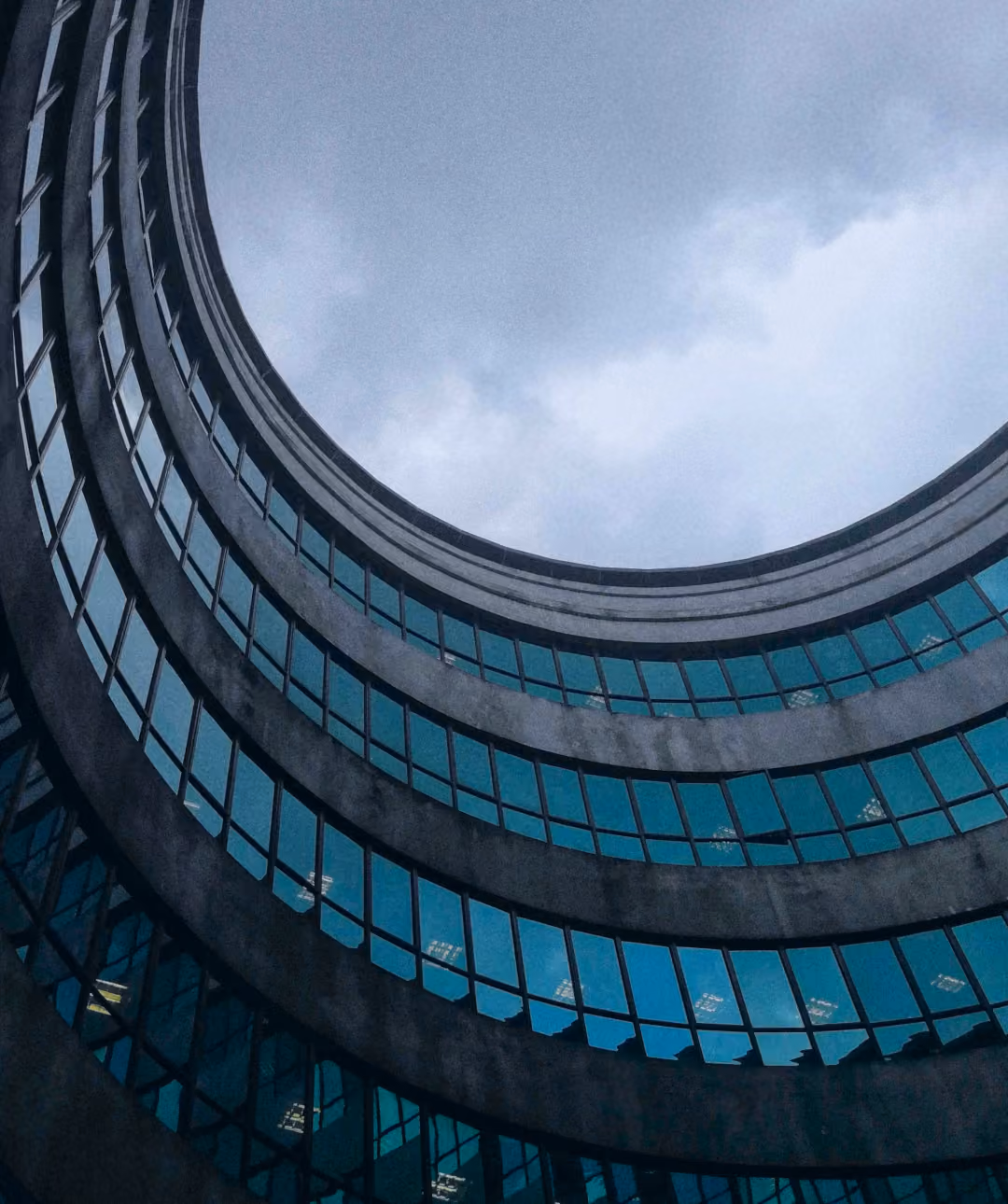 Low-angle view looking up at a circular, multi-story modern building with teal-tinted glass windows framing an overcast sky.