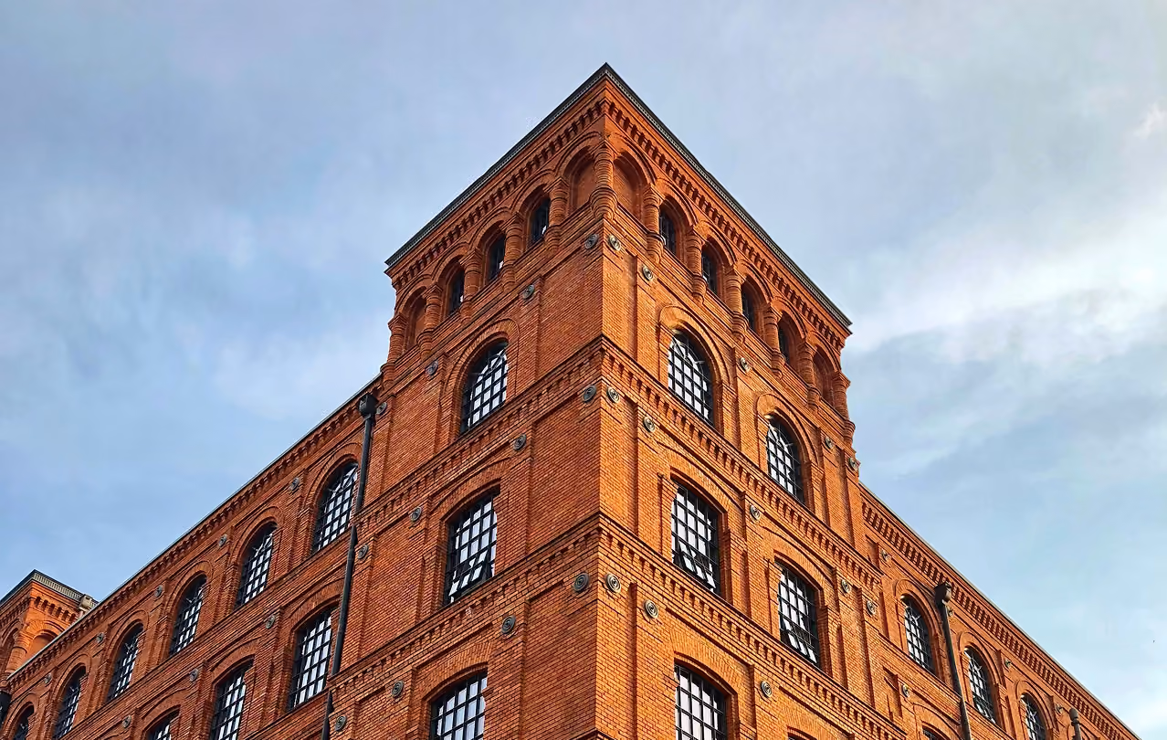Low-angle view of a multi-story red brick industrial building with arched windows and decorative brickwork against a cloudy sky.