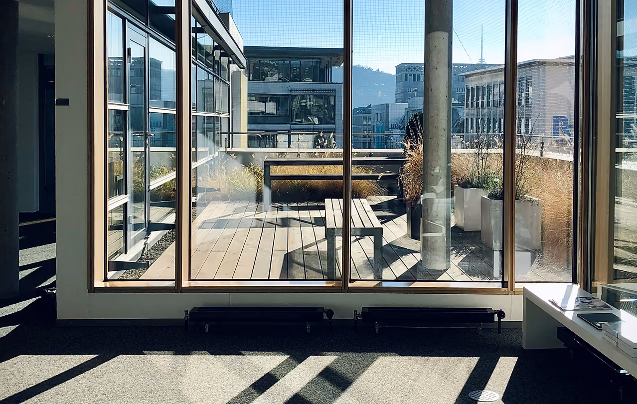 Modern office interior looking out through large glass windows onto a rooftop terrace with wooden decking, benches, and potted plants, overlooking city buildings.