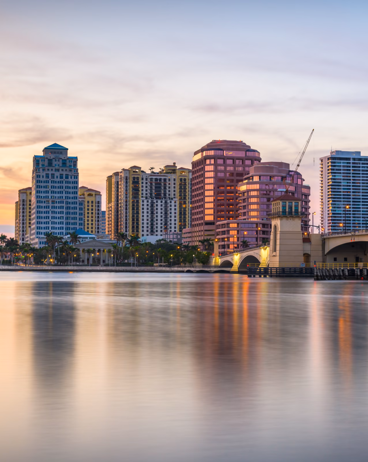 Coastal city skyline at sunset reflecting over calm water near a bridge.