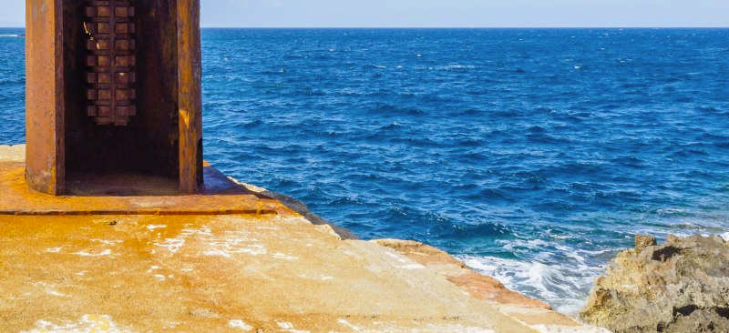 A rusty metal structure on a rocky outcrop overlooking a blue ocean