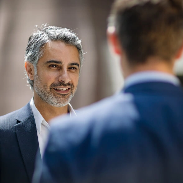 Close-up shot of a smiling, middle-aged man with salt-and-pepper hair and a beard wearing a dark suit jacket and a white dress shirt, looking towards another man whose back is partially visible in the foreground.