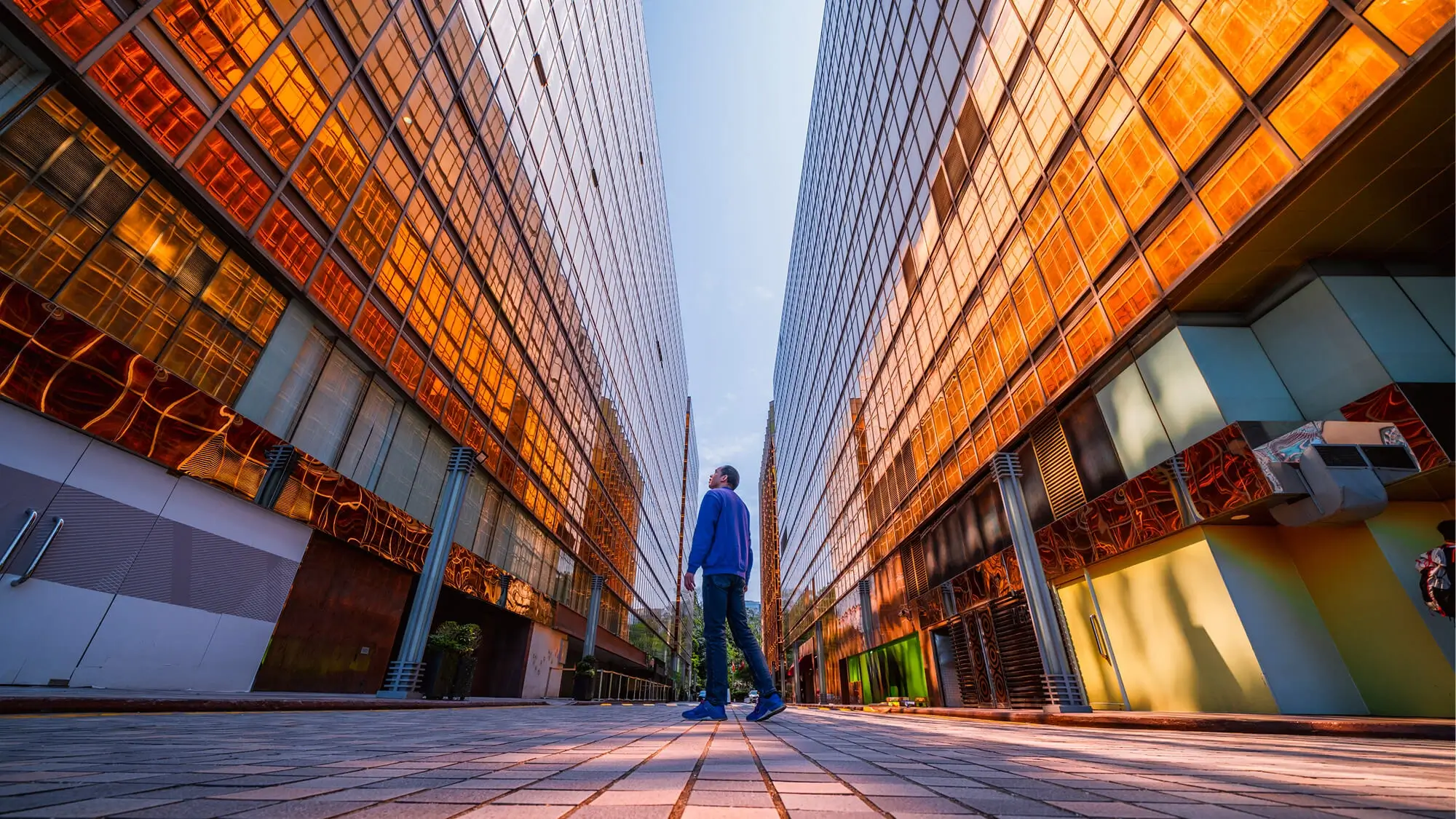 Low-angle wide shot of a man in a blue sweater standing on a paved walkway between two towering skyscrapers with reflective, golden-orange glass facades under a clear sky.