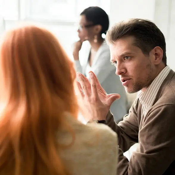 Close-up profile of a man with light brown hair gesturing with his hand while speaking intently to a woman with reddish hair, who is seen from behind in the blurred foreground of an office setting.