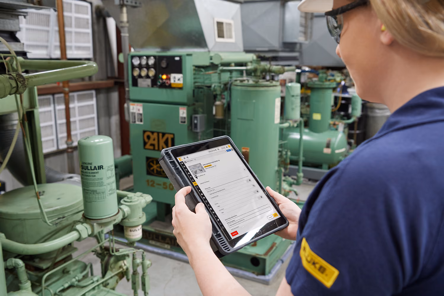 Worker in safety glasses and ear protection holds a tablet displaying a maintenance reporting app while inspecting industrial green machinery and equipment inside a facility.