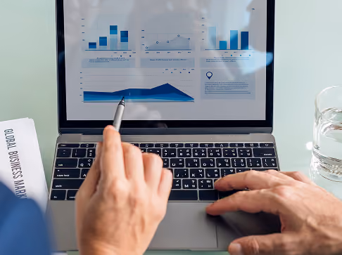 Two people viewing a laptop screen showing charts and graphs including bar charts and a line graph; one person points at the screen with a pen and the other uses the keyboard, with a 'Global Business Market' document and a glass of water nearby.