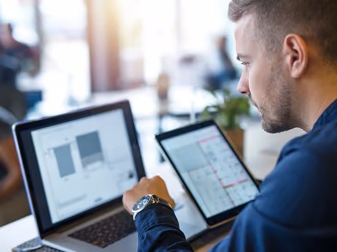 Man working on a laptop and holding a tablet displaying charts and graphs in a bright office space.