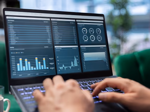 Person typing on a laptop displaying financial or business analytics dashboards with charts, graphs, and statistics on the screen.