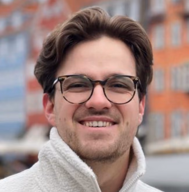 Smiling man with glasses and brown hair wearing a white fleece jacket outdoors with buildings in background.