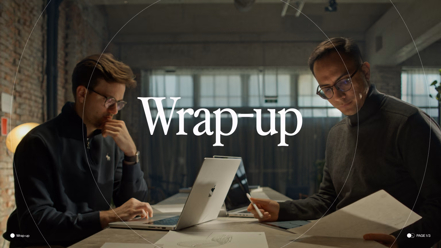 Two men with glasses working at a table with laptops and papers in an industrial-style office.