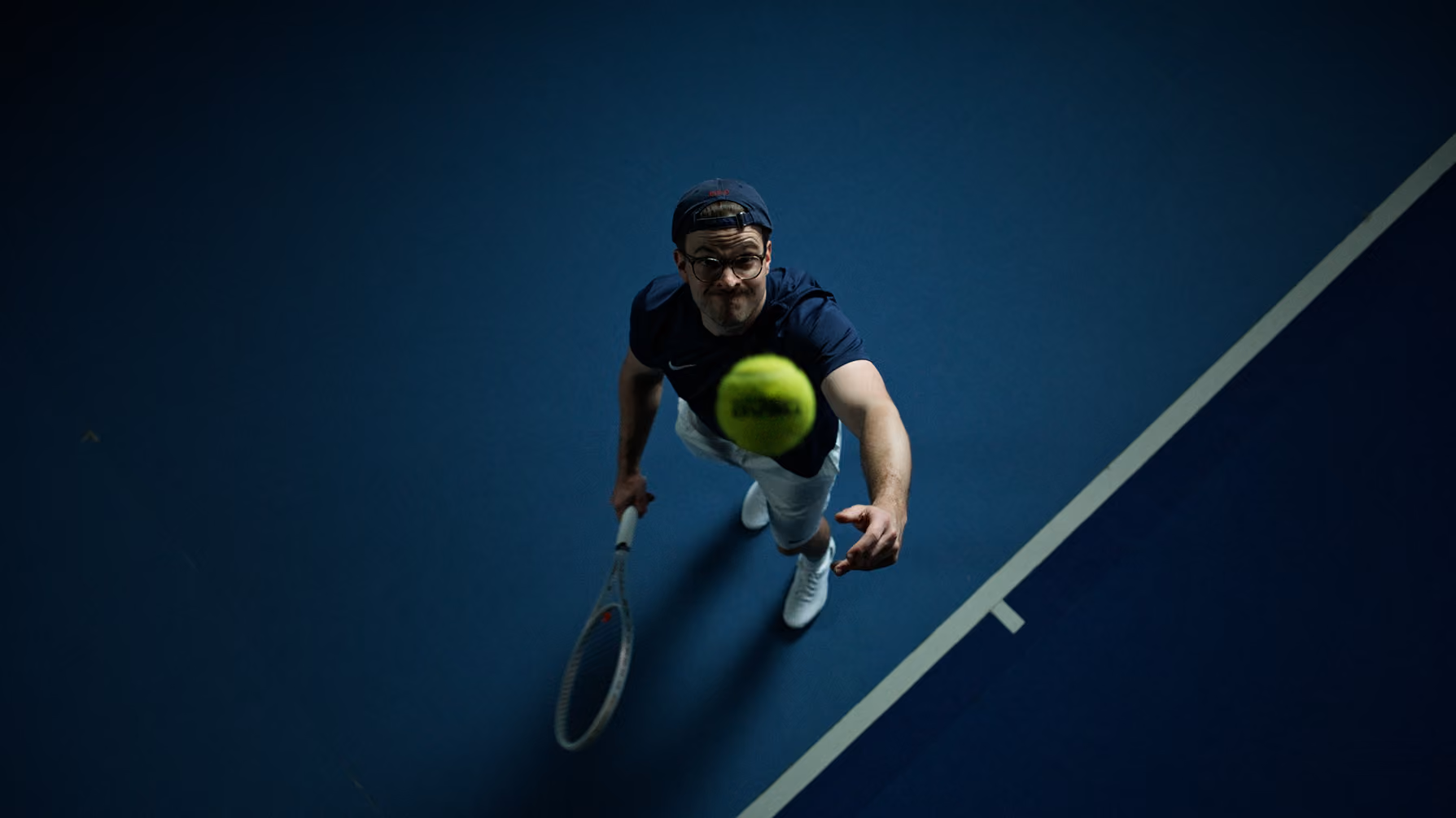 Tennis player wearing glasses and cap tossing ball up preparing to serve on an indoor court.