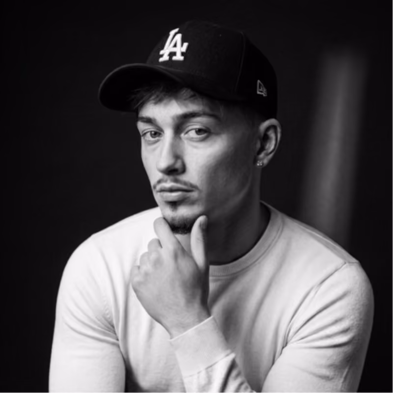 Young man in a white shirt and black LA cap with hand on chin looking at the camera in black and white.