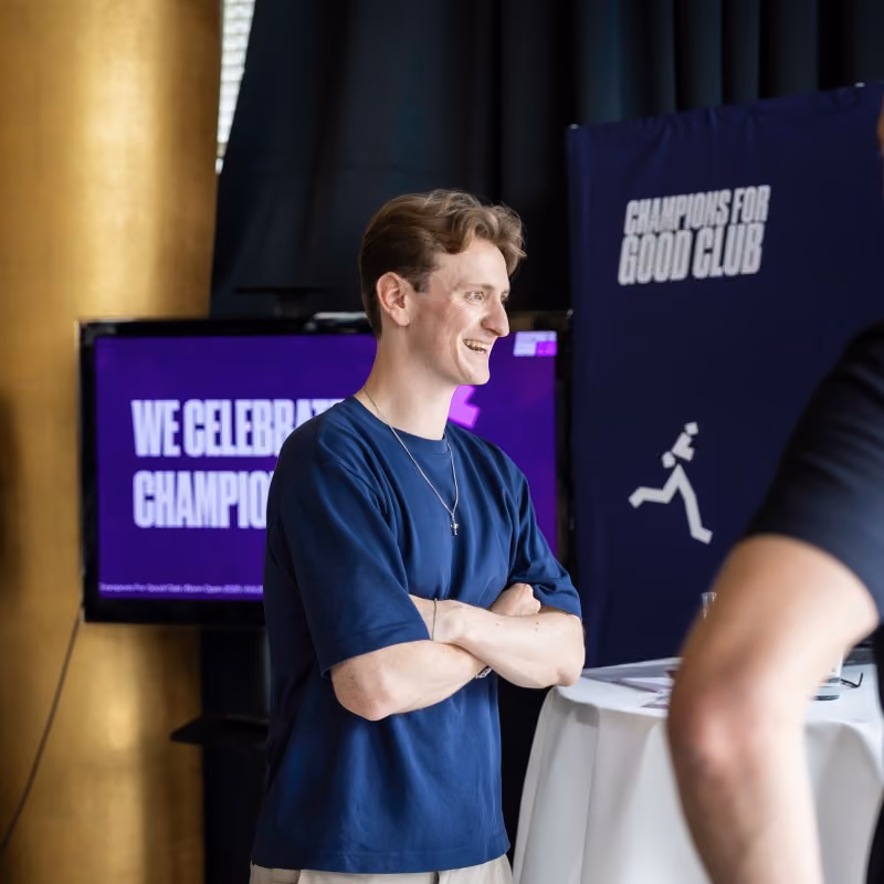 Young man in navy blue shirt smiling with arms crossed near event signs and table.