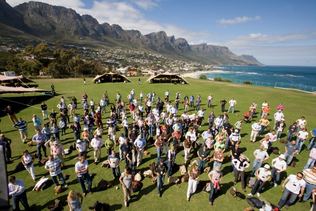 Large group of diverse people standing on green grass outdoors in a spaced formation, facing the camera, with a scenic backdrop of mountains, coastline, and blue sky.