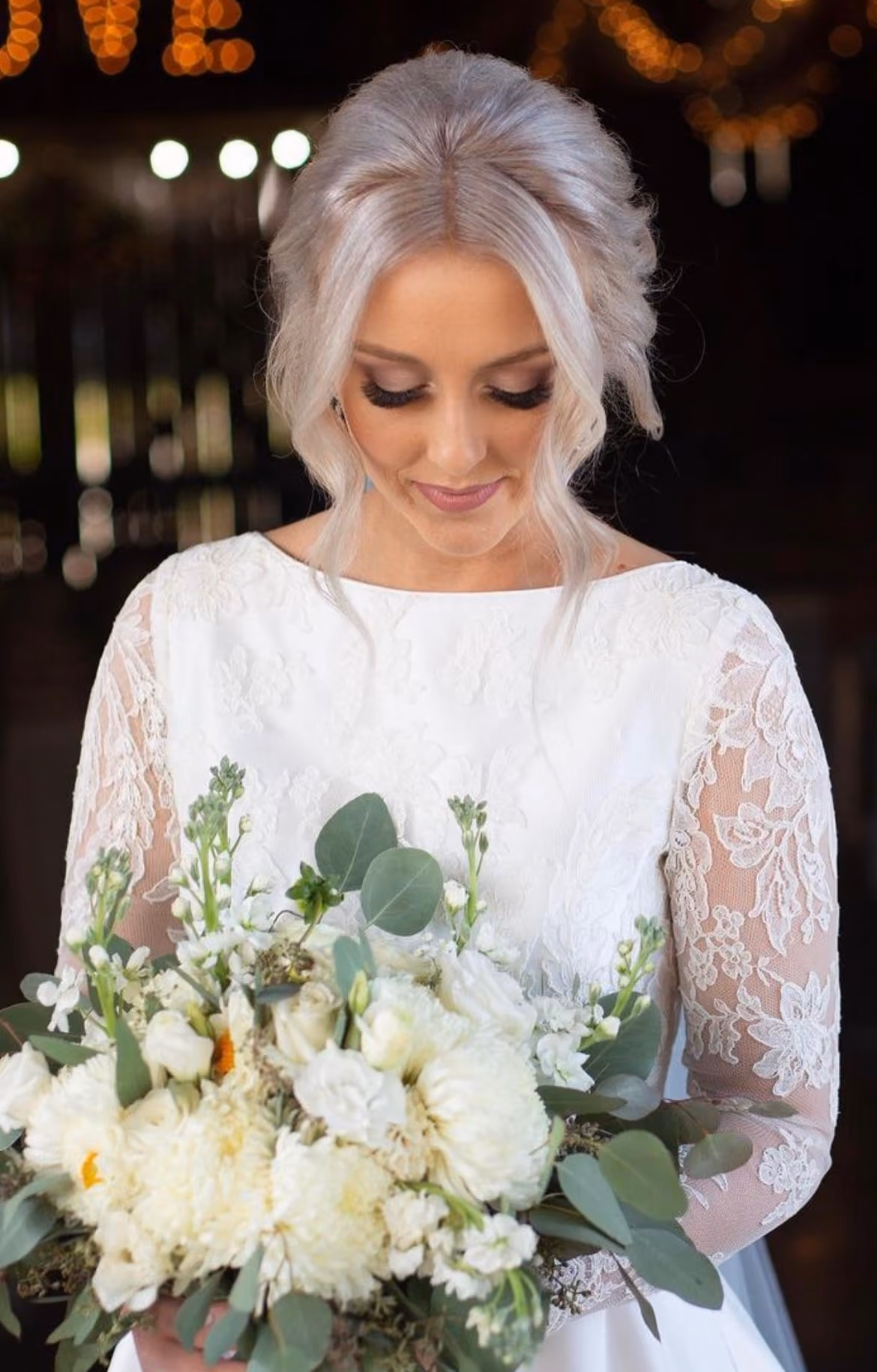 Bride with a silver-blonde updo in a lace-sleeved gown holding a white bouquet