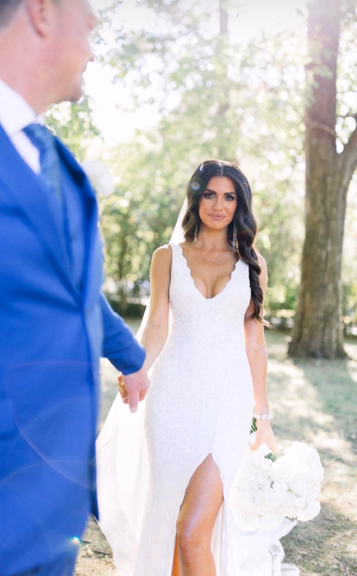 Bride in a fitted gown holding hands with her groom in a wooded setting