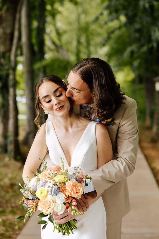 Groom kissing his bride's cheek as she holds a pastel bouquet