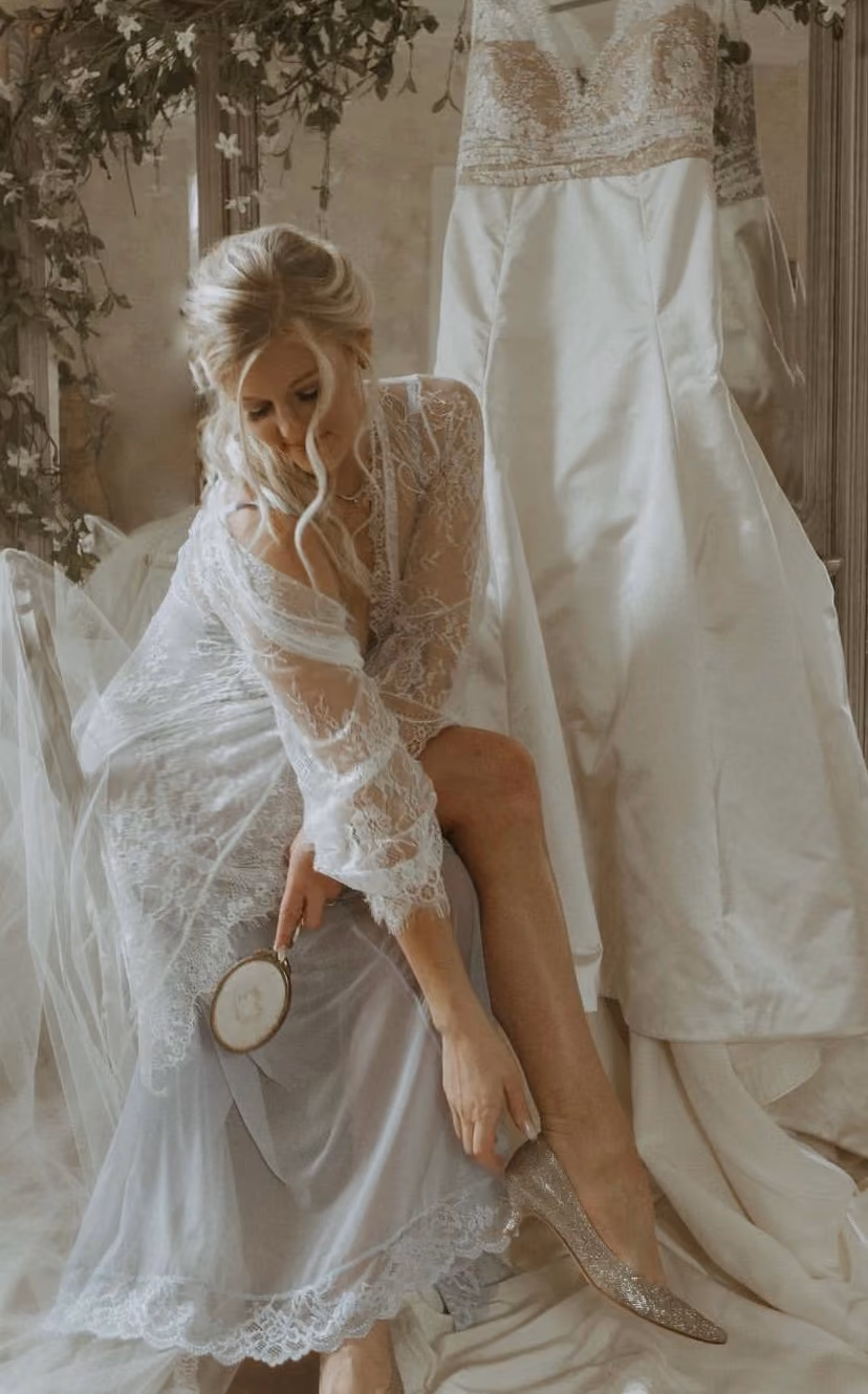 Bride in a lace robe fastening her shoe beside her hanging gown