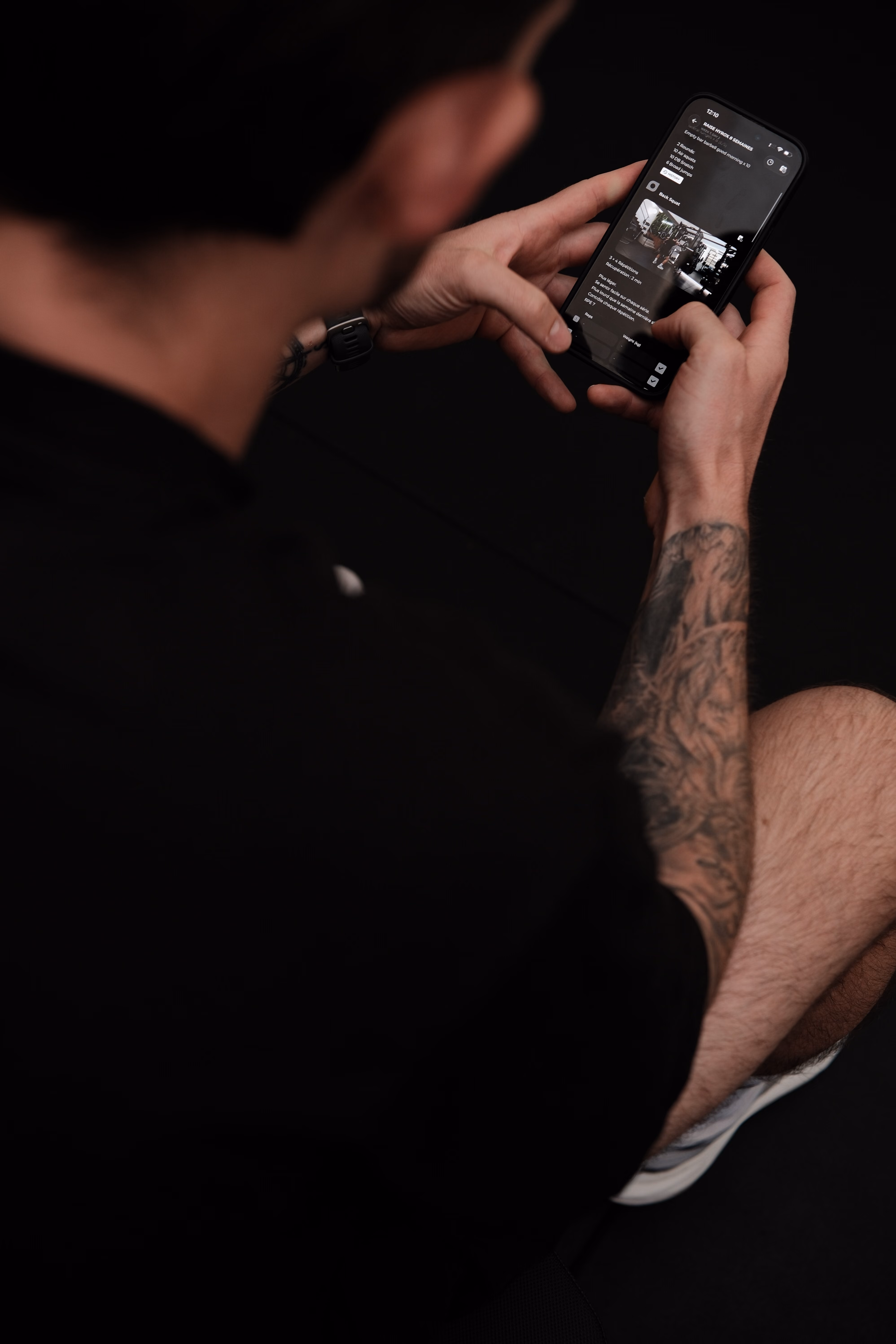 Person with tattooed arm and black shirt holding a smartphone and browsing a social media app in dark mode, against a dark background.