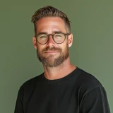 Smiling man with glasses, beard, and short hair wearing a black shirt against a plain green background.