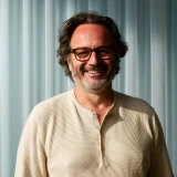 Smiling man with glasses and curly hair wearing a beige sweater standing in front of a curtain.