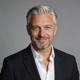 Smiling middle-aged man with gray hair and beard wearing a black jacket and white shirt against gray background.