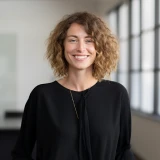 Smiling woman with curly hair wearing a black top in a bright room.