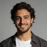 Young man with curly hair and beard smiling, wearing a black shirt over a white tee on gray background.