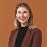 Smiling woman with shoulder-length blonde hair wearing brown blazer and black top on brown background.