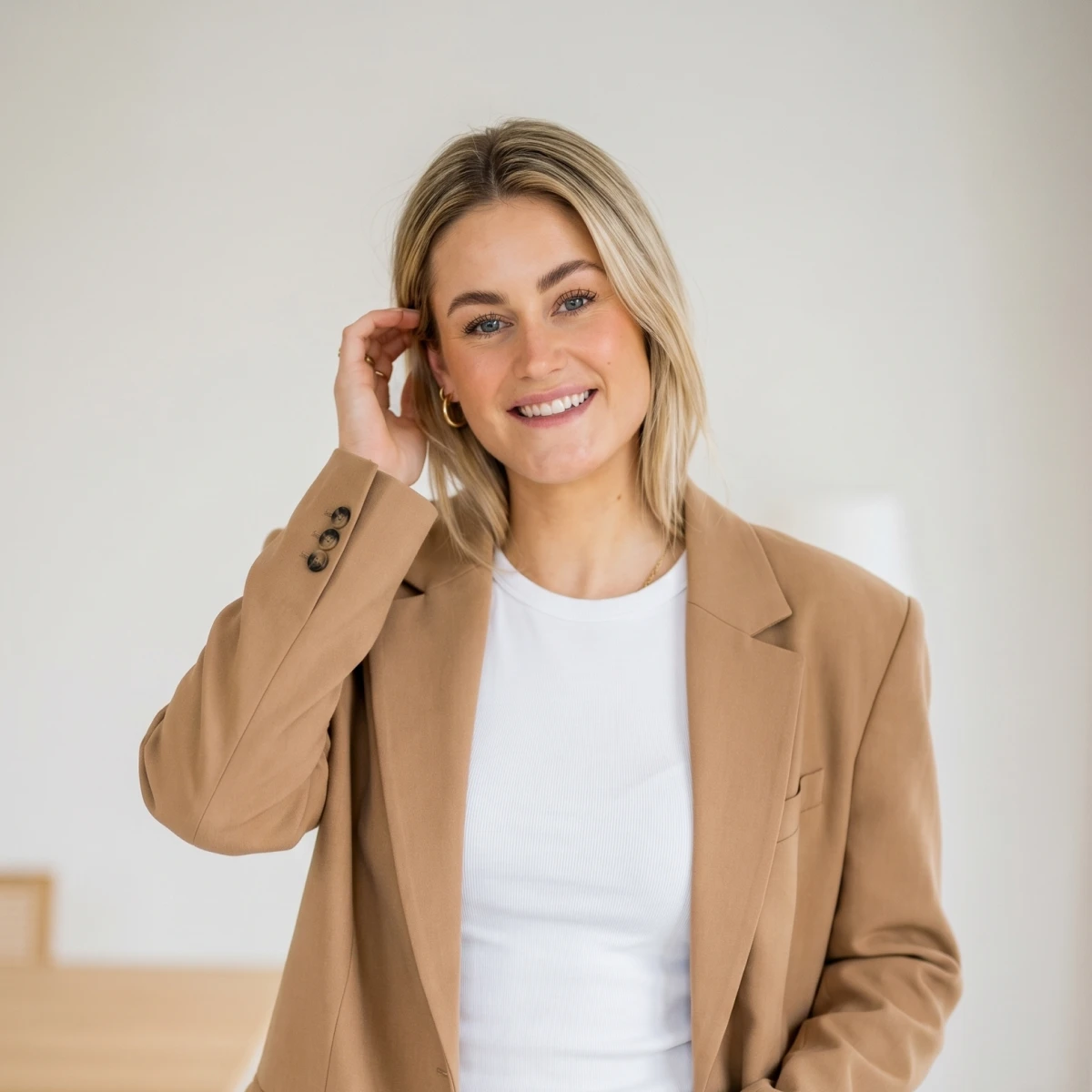 Smiling blonde woman in white shirt and tan blazer touching her hair against light background.